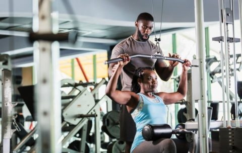 A mid adult man in his 30s working as a fitness instructor in a gym, helping a mature woman in her 50s strengthening her arms on exercise equipment. They are both African-American.