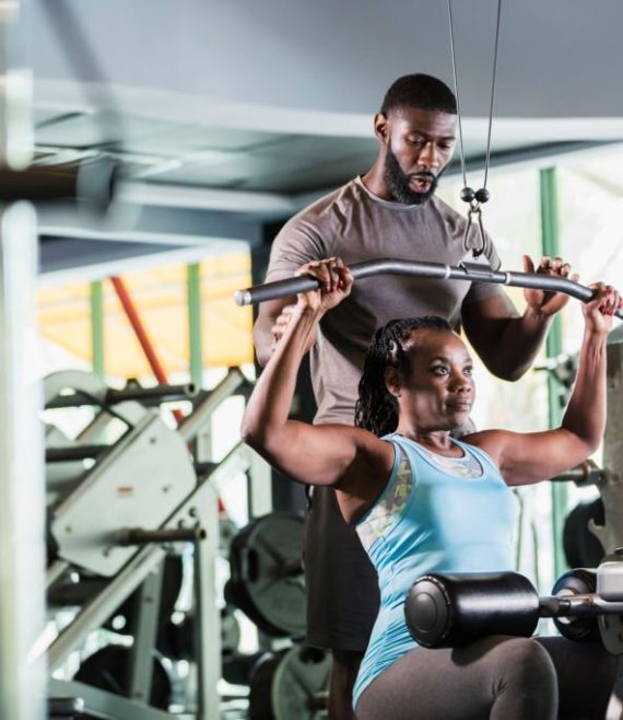 A mid adult man in his 30s working as a fitness instructor in a gym, helping a mature woman in her 50s strengthening her arms on exercise equipment. They are both African-American.
